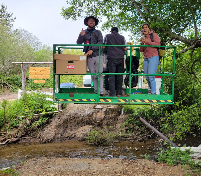 Two men and a woman ride in green bucket lift over the creek.