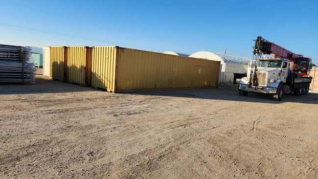 Three shipping containers sit in front of greenhouses. A truck with a folded crane is parked beside them.