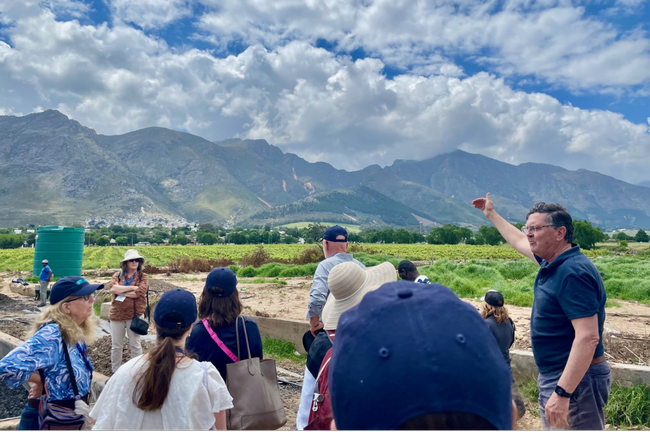 A man gestures toward a green crop field as tour participants listen to him speak.