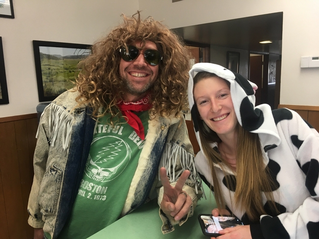 Ryan, wearing a curly, long blond wig, dark sunglasses, green Grateful Dead shirt and jean jacket trimmed with leather fringe, makes a peace sign with his left hand. Samantha leans over a counter toward him wearing a black and white cow costume.