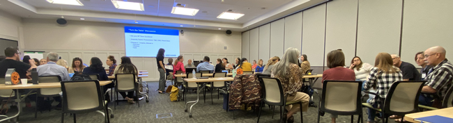 Dozens of people sit at conference room tables. PowerPoint screen reads: 