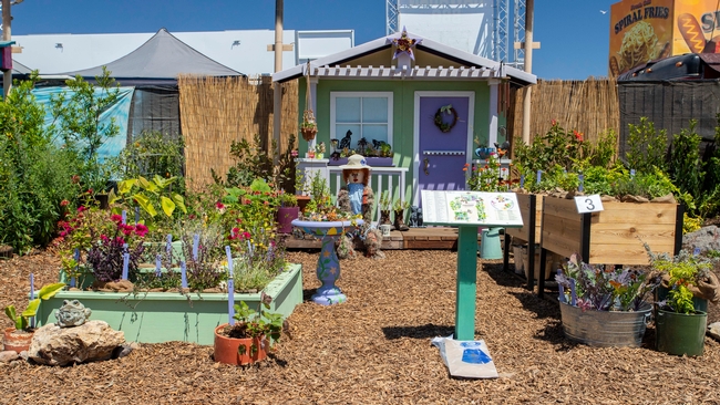 Plants in containers in front of a little house. A guide identifying the plant varieties sit atop a stand.