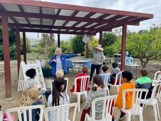 Two middle aged women stand in front of a crowd of seated children, teaching them about food.