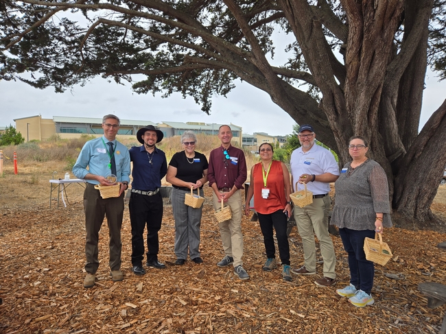 Seven people stand under a tree, five are holding baskets.