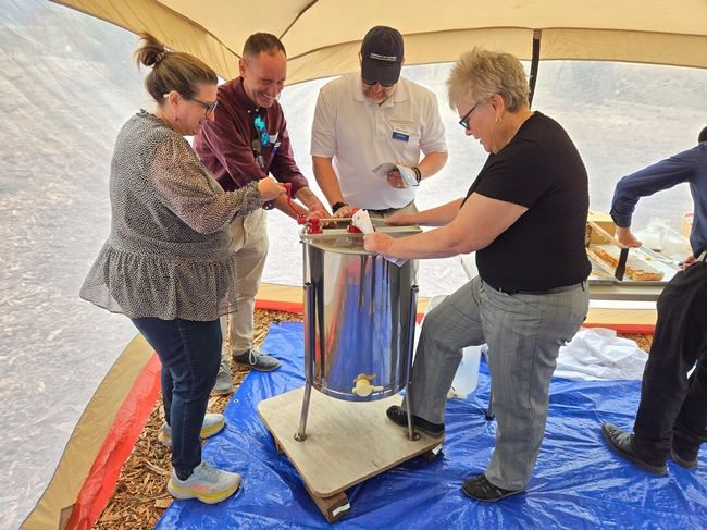 Four people are gathered around a shiny, silver-colored pot. Elizabeth turns the hand crank as the others watch.