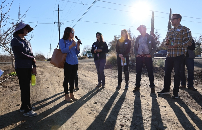 Hope, surrounded by 6 people standing on a dirt road, speaks into a mic.