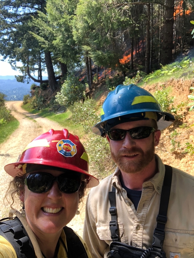 Lenya and Jeff are outdoors wearing their protective red and blue helmets as a prescribed fire burns among the trees behind them.