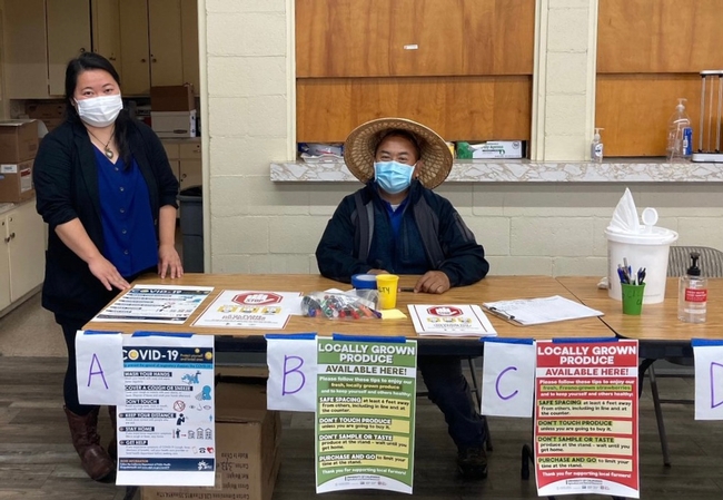 Two UCCE Hmong small farms representatives sit behind a table displaying COVID-19 signs.