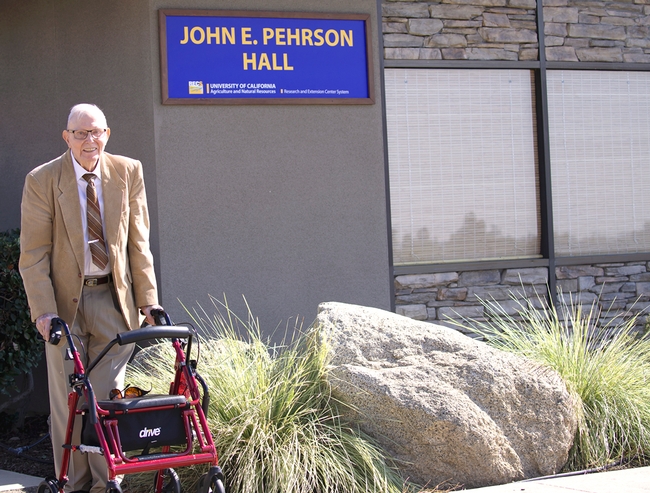 John Pehrson, standing with a walker, poses beside the building sign bearing his name.