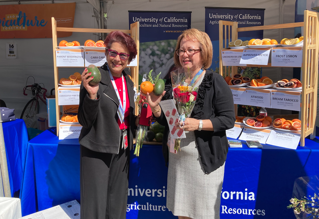 Senators Anna Caballero and Maria Elen Durazo each hold citrus, avocados and a bouquet of flowers while standing in front of the ANR booth.