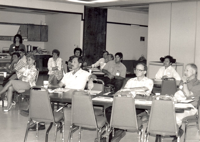 Black and white photo of Bill Weir and others sitting at long tables listening to a presentation.