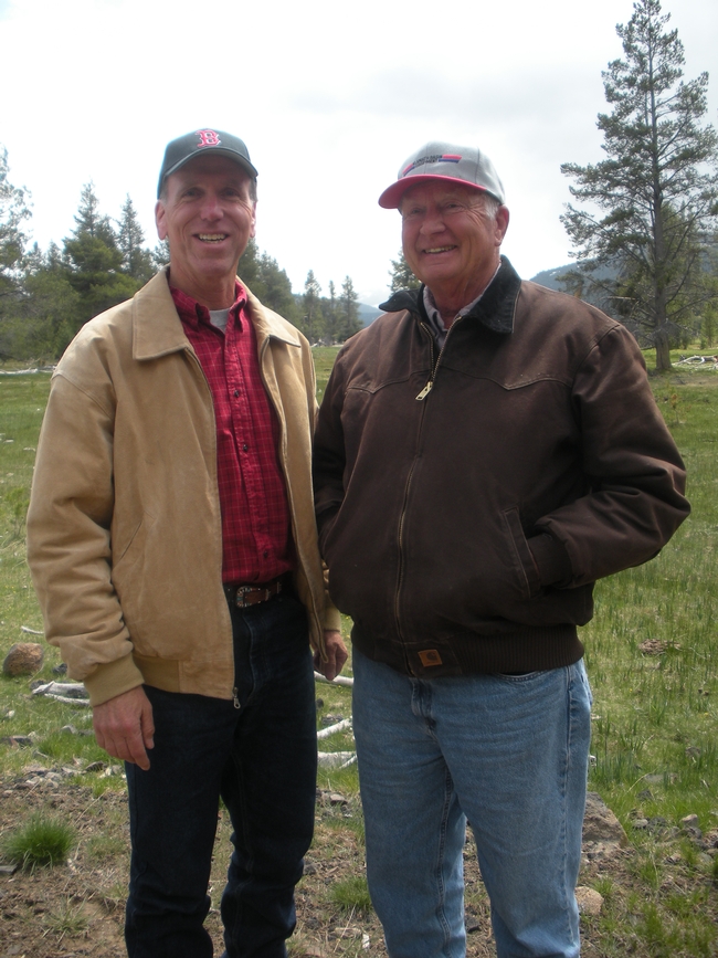 Two men wearing jackets and ball caps stand side by side outdoors.