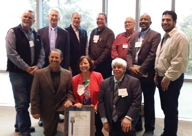 Duncan and two other people kneel beside a plaque. Seven men stand behind them.