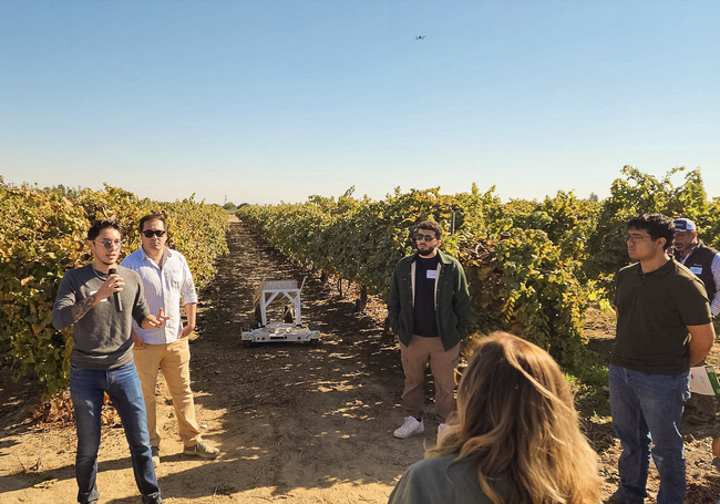 A man standing in a vineyard speaks into a mic as people listen. The Rover robot rolls  between rows of grapevines.