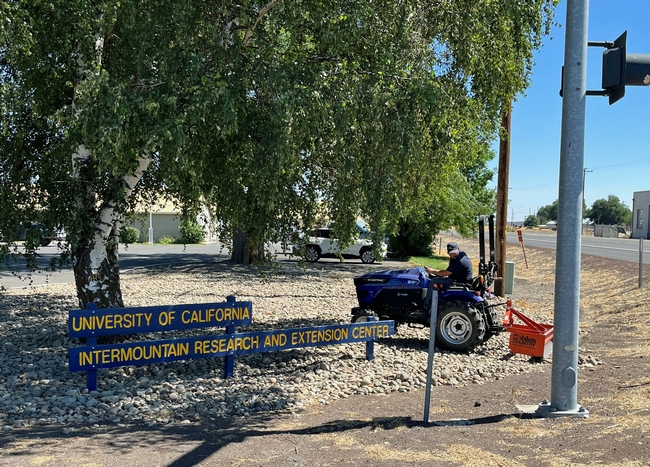 A man drives a small blue tractor over rocks around the University of California Intermountain Research and Extension Center sign.