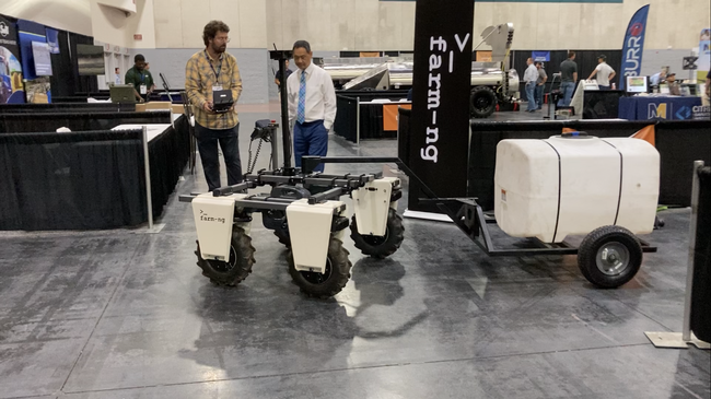 In an exhibit hall, Rublee uses handheld control to move a 4-wheeled robot to pull a water tank on a trailer.