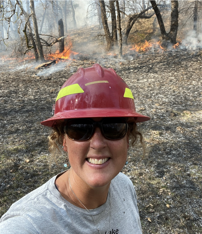 Selfie of Lenya wearing a hart hat with flaming vegetation in the background.