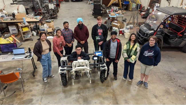 Nine people stand around the Huskerbot in a workshop.