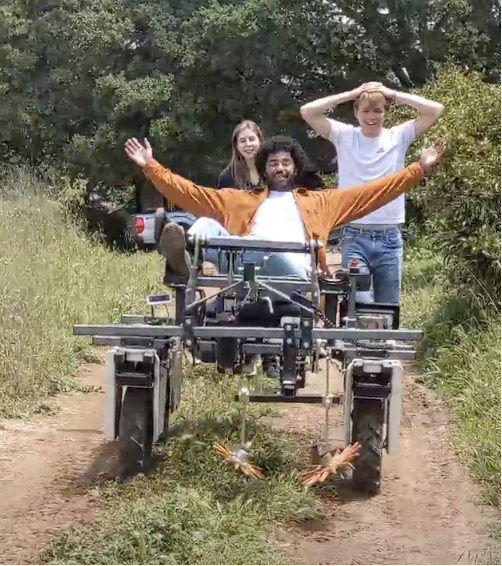 One man reclines with outstretched arms atop the robot with two people standing behind the machine.