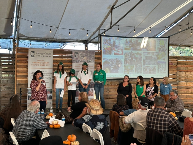 A woman speaking into a mic introduces the 4-H members, who are wearing green and white uniforms standing on a stage. Audience members sit in the foreground.