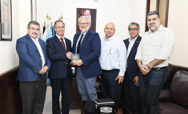 Six men stand. The University of Jordan President Nathir Obeidat hands Brent a plaque and shakes his hand.