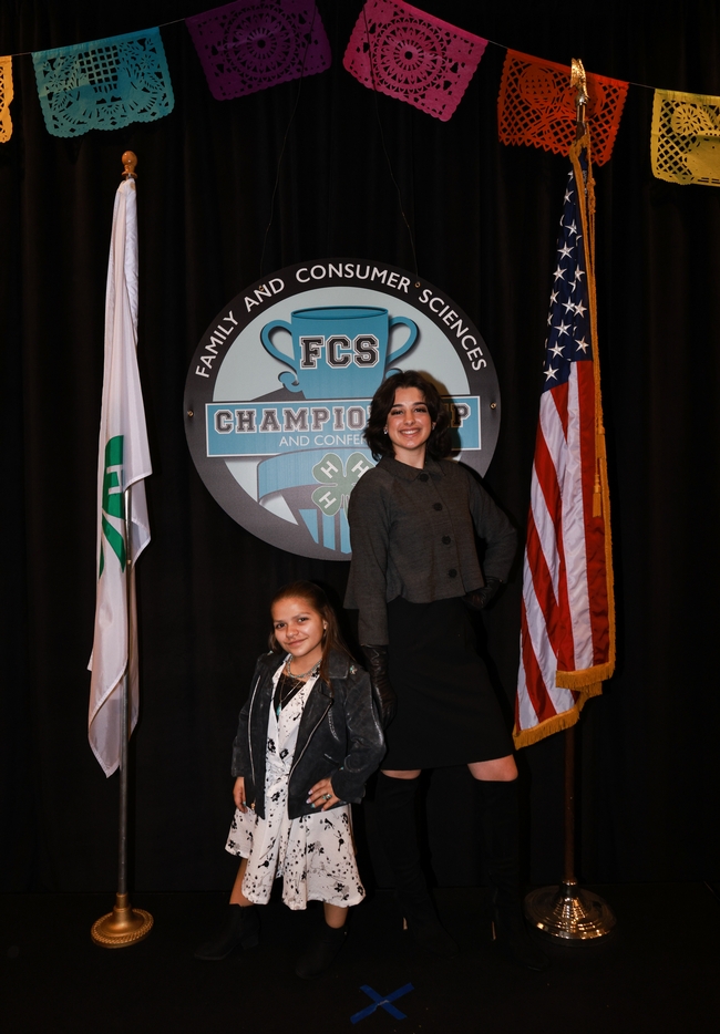 Lily and Olivia pose in their winning outfits in front of FCS Championship sign, between 4-H flag and U.S. flag