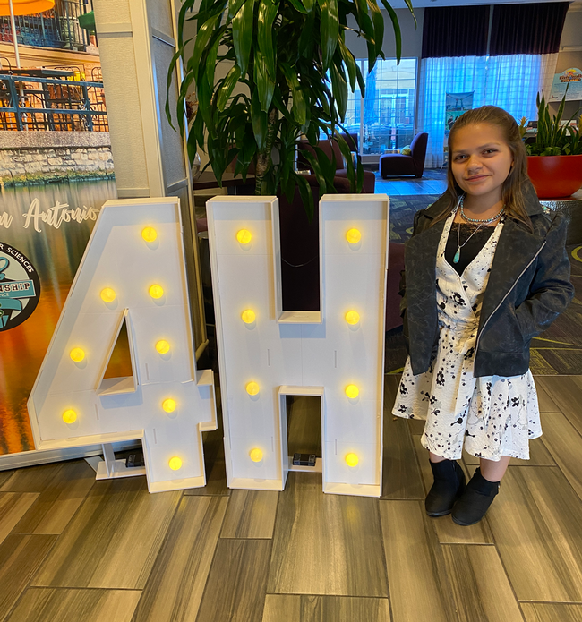 Lily wearing a faux leather jacket and wrap dress, standing next to a lighted 4-H sign