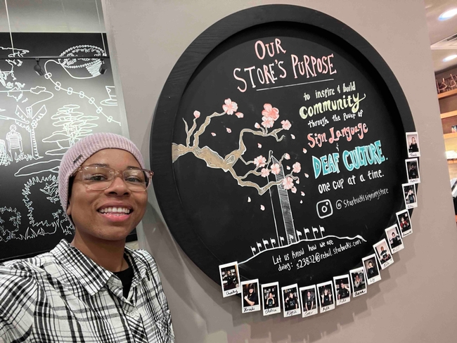 Eve in front of round sign saying Our Store's purpose: to inspire & build community through the power of sign language & deaf culture. One cup at a time.