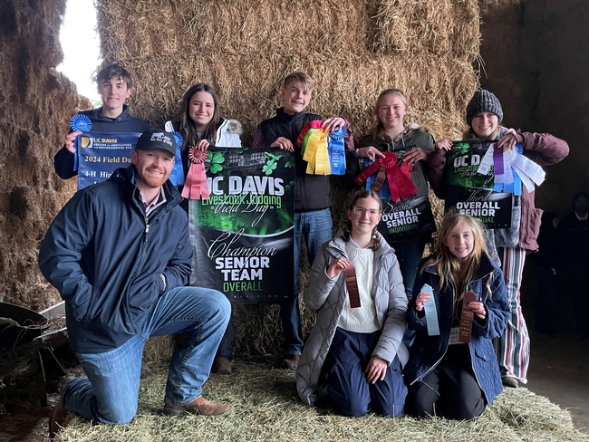 Against a backdrop of hay bales, 5 youth hold ribbons and posters of their contest placing. Kneeling in front of them is an adult coach and 2 youth holding ribbons.