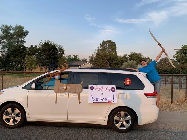 White van decorated with an image of a deer and sign saying Archery is Awesome. Youth on back holding a bow.