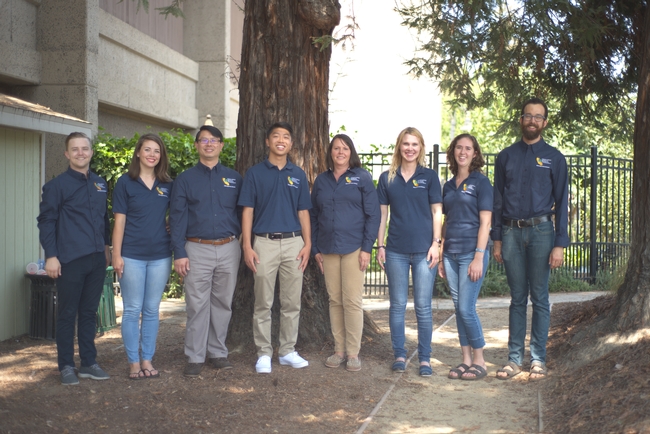 8 people in dark blue shirts standing in a row under a large tree