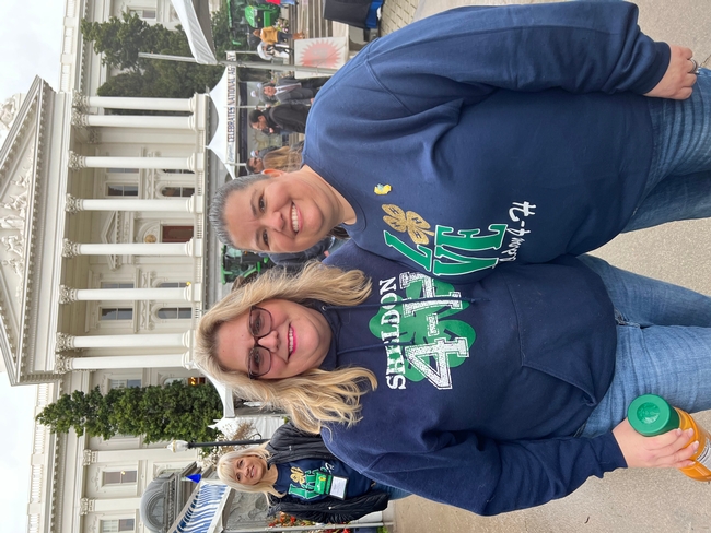 The image shows two women posing together, smiling. Both are wearing Sheldon 4-H tops and jeans.