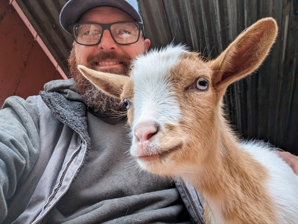 A man with a hat, glasses and a beard next to a brown and white goat.