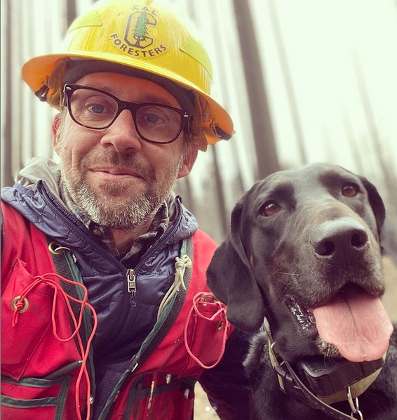 Ryan Tompkins wearing a yellow hard hat and red jacket with his black labrador retriever