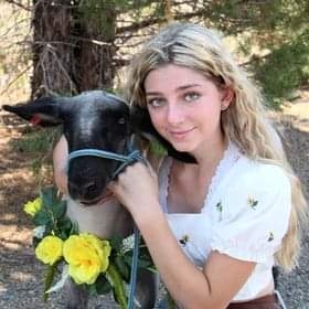 Blonde-haired girl poses next to lamb that is wearing a wreath of yellow roses around its neck.