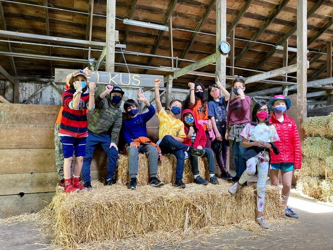 Ten students sit on straw bales raising their forearms to show off the bracelets they made from sheep wool.