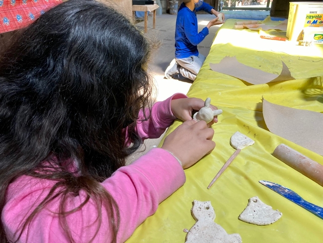 A girls with long dark, wavy hair and a pink jacket uses her fingers to form a bunny out of clay.