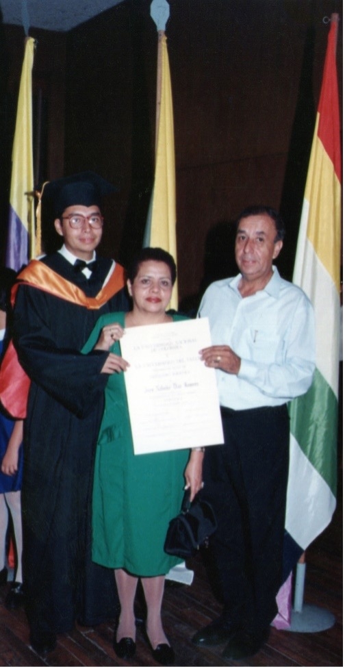 A young man wearing a cap and gown and glasses, poses with his mother and father while holding up his college degree.