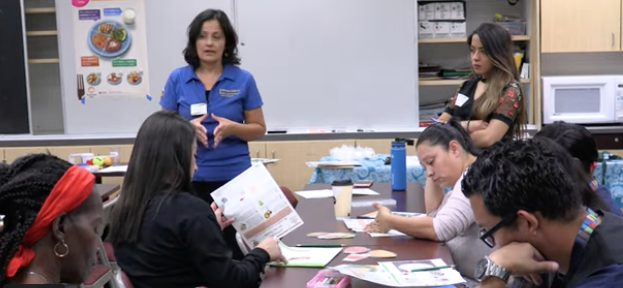 Wilshire, wearing a blue polo shirt, speaks to a classroom of students seated at tables. Behind her is a poster showing MyPlate of food.