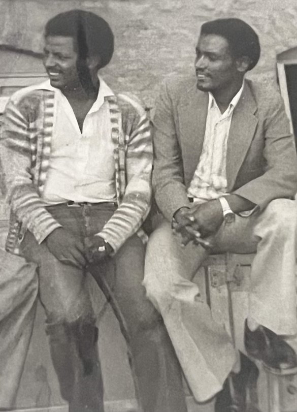 Two men sitting against a wall, looking off camera.