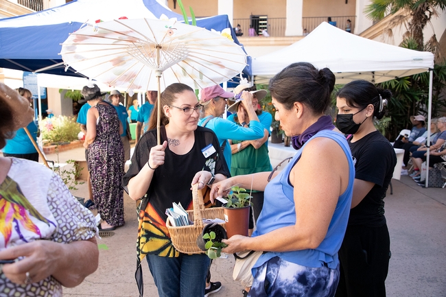 One woman is holding a basket with pamphlets as another woman reaches into the basket with one hand, while holding plants in the other.