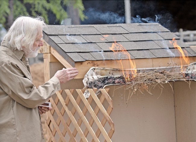 Steve Quarles gestures toward flames rising from dry pine needles and leafy debris in a melting plastic rain gutter affixed to a structure with a shingled roof.