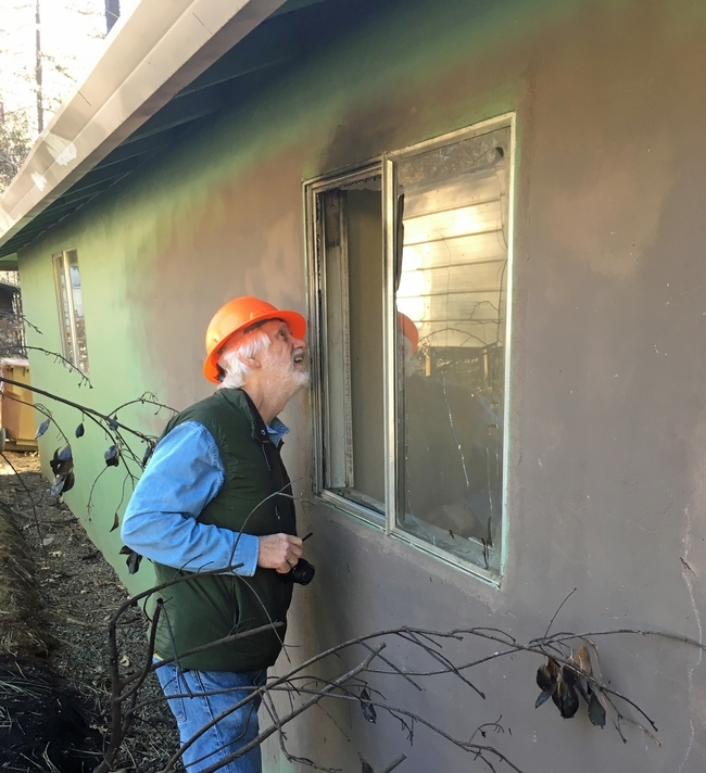 Steve Quarles, wearing an orange hard hat, peers into a shattered window of a garage. The exterior of the pistachio green one-story building is gray around the broken window.