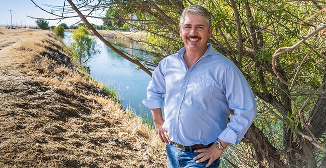 Josh Viers stands beside a small tree on a bank beside an irrigation canal in a rural setting.