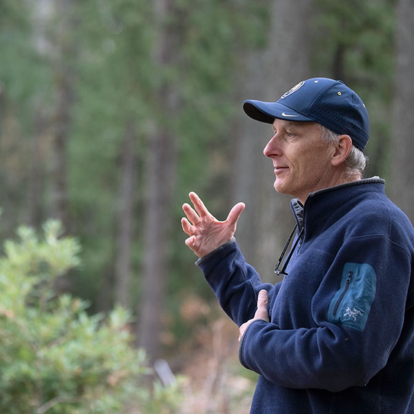Wearing a navy blue cap and jacket, John Battles speaks while standing in the forest.
