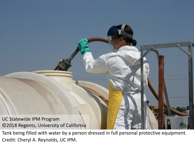 A person filling a tank with water while wearing chemical-resistant gloves, coveralls, chemical-resistant apron, and a face shield over safety glasses.