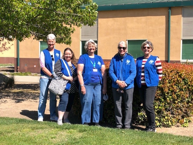 Five smiling women in front of a garden wearing blue vests.