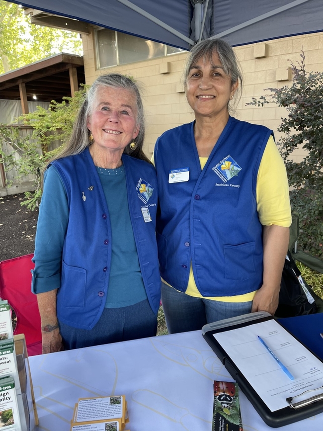 Two women wearing blue vests and smiling while staffing a booth.