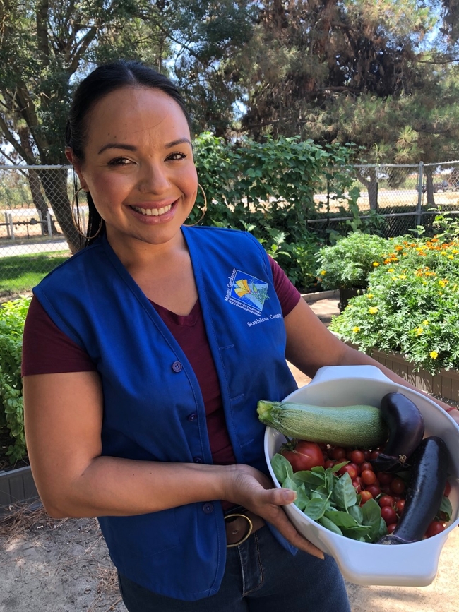 Woman wearing Master Gardener vests holds a basket of colorful summer vegetables.