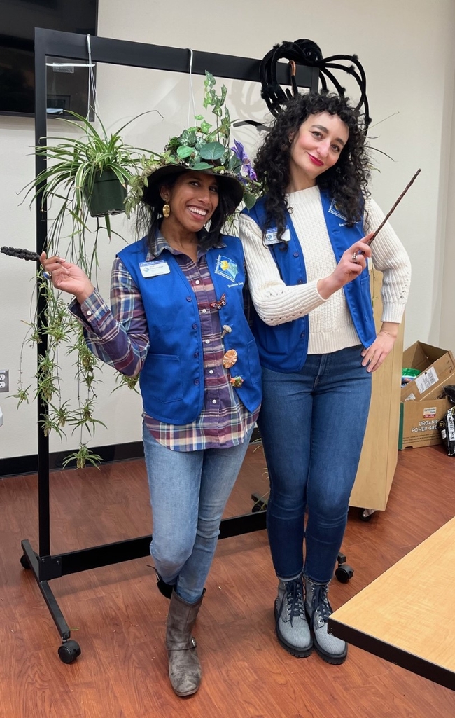 Two women wearing Master Gardener vests smiling in front of a spider plant and fake, oversized black spider.
